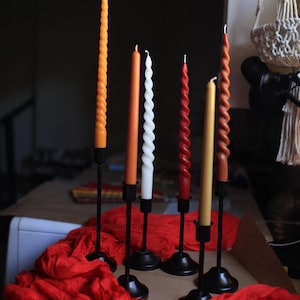 K&ouml;nnte beinhalten: Sechs Stabkerzen in verschiedenen Farben wie Orange, Wei&szlig;, Rot und Gelb, einige mit gedrehter Spiralstruktur und andere glatt. Jede Kerze steht aufrecht in einem schwarzen Metallkerzenhalter mit rundem Fu&szlig; und einem d&uuml;nnen Stiel.