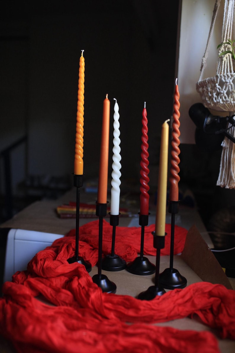 K&ouml;nnte beinhalten: Sechs Stabkerzen in verschiedenen Farben wie Orange, Wei&szlig;, Rot und Gelb, einige mit gedrehter Spiralstruktur und andere glatt. Jede Kerze steht aufrecht in einem schwarzen Metallkerzenhalter mit rundem Fu&szlig; und einem d&uuml;nnen Stiel.