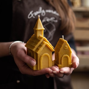 Puede incluir: Dos velas amarillas de cera de abeja con forma de iglesias. La vela más grande tiene un campanario y un tejado detallado, mientras que la más pequeña tiene forma de casa sencilla. Ambas tienen una mecha y se sostienen en las manos.