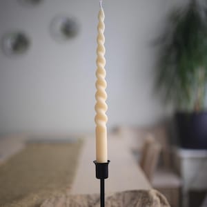 K&ouml;nnte beinhalten: Eine lange, cremefarbene, gedrehte Stabkerze mit spiralf&ouml;rmigem Design. Die Kerze steht aufrecht in einem schlichten, mattschwarzen Metallkerzenhalter mit rundem Fu&szlig; und schlankem Stiel und besitzt einen einzelnen wei&szlig;en Docht.