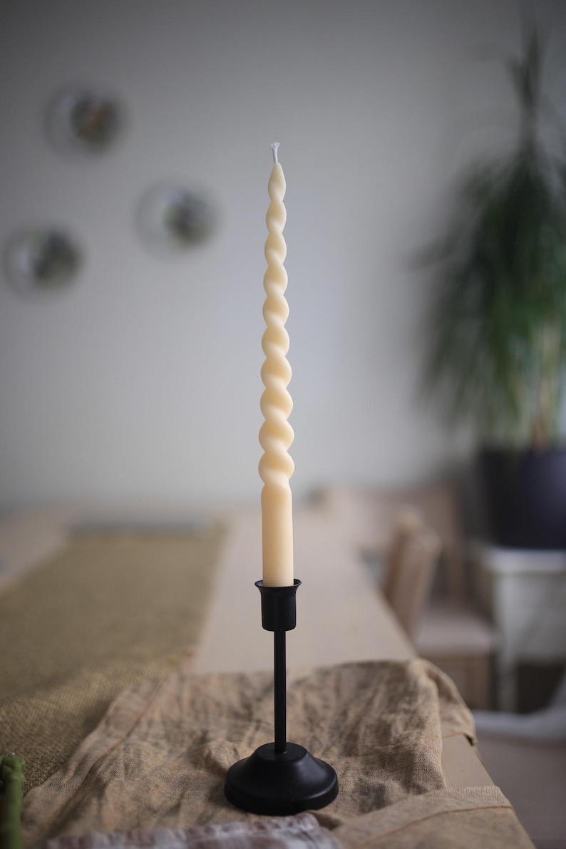 K&ouml;nnte beinhalten: Eine lange, cremefarbene, gedrehte Stabkerze mit spiralf&ouml;rmigem Design. Die Kerze steht aufrecht in einem schlichten, mattschwarzen Metallkerzenhalter mit rundem Fu&szlig; und schlankem Stiel und besitzt einen einzelnen wei&szlig;en Docht.