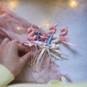 Könnte beinhalten: Eine Sammlung von ziffernförmigen Kerzen auf Holzstäbchen, die die Zahlen eins und zwei in Rosa-, Blau- und Weißtönen zeigen. Jede Kerze ist mit einer kleinen Satinschleife verziert, die um die Basis des Holzstäbchens gebunden ist.