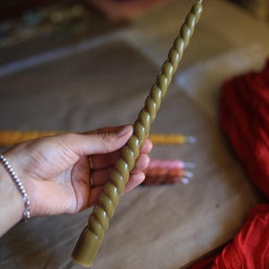K&ouml;nnte beinhalten: Eine lange, schlanke Stabkerze in ged&auml;mpftem Olivgr&uuml;n mit einer durchgehenden, gedrehten, seilartigen Textur &uuml;ber die gesamte L&auml;nge und einem einzelnen wei&szlig;en Docht am spitzen oberen Ende, geeignet f&uuml;r handels&uuml;bliche Kerzenhalter.