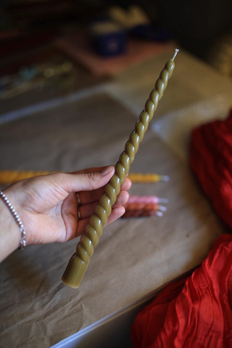 K&ouml;nnte beinhalten: Eine lange, schlanke Stabkerze in ged&auml;mpftem Olivgr&uuml;n mit einer durchgehenden, gedrehten, seilartigen Textur &uuml;ber die gesamte L&auml;nge und einem einzelnen wei&szlig;en Docht am spitzen oberen Ende, geeignet f&uuml;r handels&uuml;bliche Kerzenhalter.
