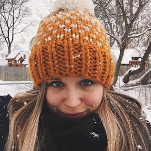 May include: A close-up of a person wearing a knitted orange and white beanie with a white pom-pom on top. The beanie is covered in snow.