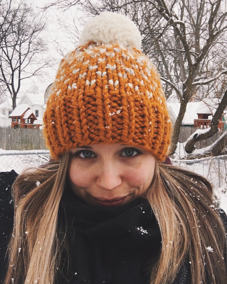 May include: A close-up of a person wearing a knitted orange and white beanie with a white pom-pom on top. The beanie is covered in snow.
