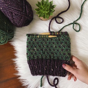 May include: A green and purple knitted hat with a speckled pattern is being worked on with knitting needles. The hat is on a white fluffy surface with green and purple yarn.
