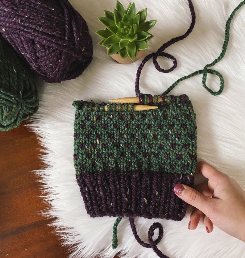 May include: A green and purple knitted hat with a speckled pattern is being worked on with knitting needles. The hat is on a white fluffy surface with green and purple yarn.