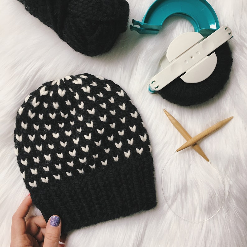 May include: A black and white knitted hat with a geometric pattern. The hat is made with a loom and is sitting on a white fluffy surface.