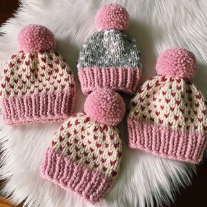 May include: Three pink and white knit hats with pom poms. The hats have a heart pattern and are made with a soft yarn.