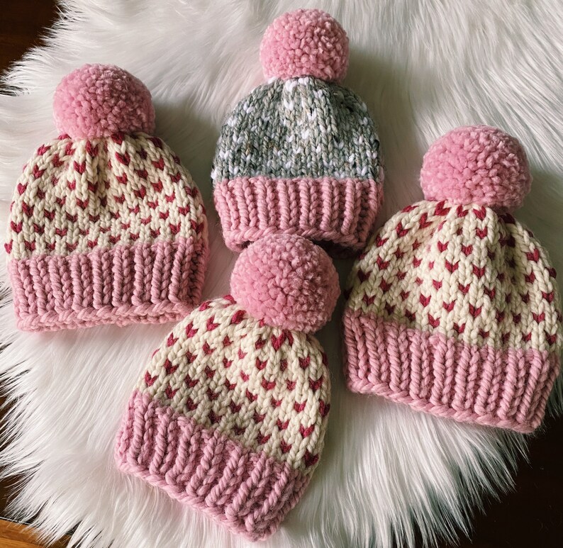 May include: Three pink and white knit hats with pom poms. The hats have a heart pattern and are made with a soft yarn.