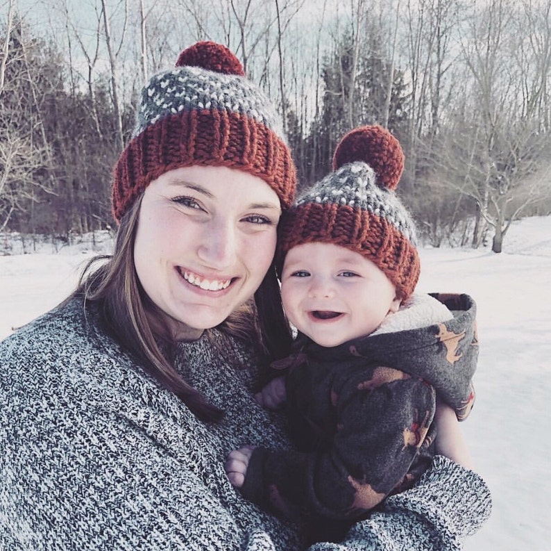 May include: A woman and a baby wearing matching red and gray knit hats with pom-poms. The woman is wearing a gray sweater and the baby is wearing a brown and green patterned jacket.