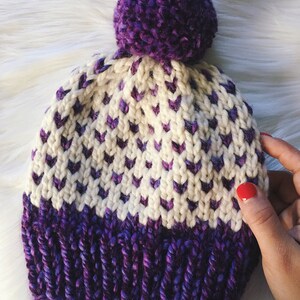 May include: A white and purple knitted hat with a purple pom-pom on top. The hat has a chevron pattern and a solid purple band at the bottom.