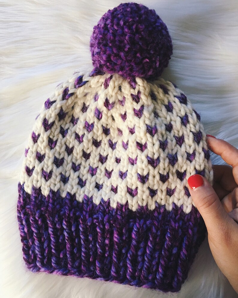 May include: A white and purple knitted hat with a purple pom-pom on top. The hat has a chevron pattern and a solid purple band at the bottom.