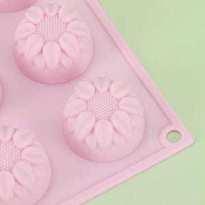 Puede incluir: Primer plano de un molde de silicona rosa con seis cavidades en forma de girasol. Cada flor tiene detalles de pétalos y un centro texturizado. El molde está sobre una superficie rosa con una esquina redondeada y un agujero.