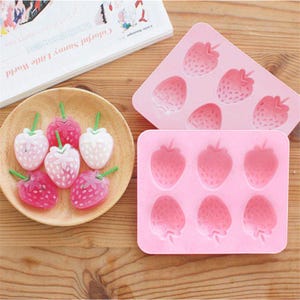 May include: A wooden plate holds six strawberry-shaped candies with green stems. Two pink silicone molds, one with six strawberry shapes and the other with eight, sit on a wooden surface. A book is in the background.