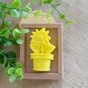 May include: A yellow soap bar with a raised design of a sunflower in a pot. The soap is in a brown frame.