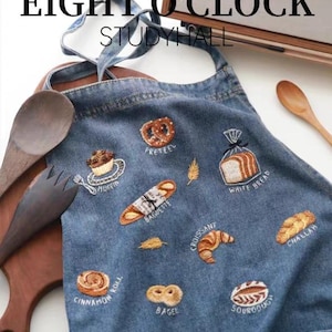 May include: Blue denim apron featuring embroidered bread and pastry designs: pretzel, muffin, baguette, croissant, bagel, and white bread. The apron has the text "EIGHT O'CLOCK STUDY HALL" at the top.