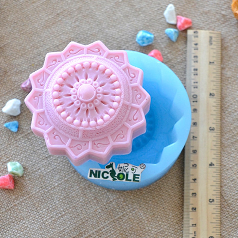 Puede incluir: Un molde de silicona rosa con un dise&ntilde;o detallado de un objeto circular con forma de estrella. El molde est&aacute; sentado sobre una base de silicona azul con la palabra "NICOLE" impresa en ella.