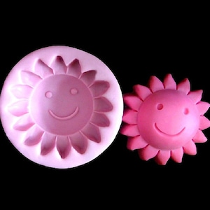 Puede incluir: Molde de silicona rosa para hornear un pastel con forma de sol sonriente. El molde se muestra junto a un pastel rosa con forma de sol sonriente.