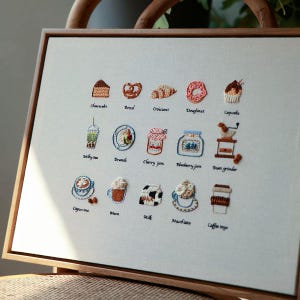 May include: Embroidered artwork featuring various food and drink items, including a cupcake, croissant, and coffee. The artwork is framed in a wooden frame and includes the text "Cheesecake", "Brunch", "Cappuccino", and more.