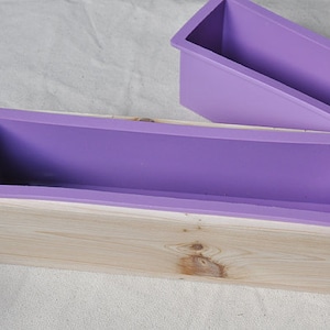 Op de afbeelding: Een paarse siliconen mal voor het maken van zeepstaven zit in een houten doos. De mal is rechthoekig en heeft een glad oppervlak.