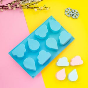 May include: A light blue silicone mould featuring cloud and teardrop shapes. The mould sits on a pink and yellow background, with two snowflake decorations and some cloud and teardrop shaped objects in pink and light blue.