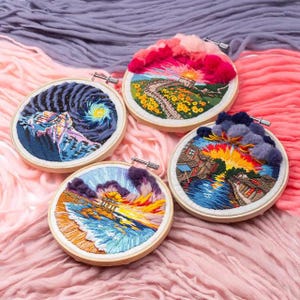 May include: Four round embroidery hoops with landscape designs. Each features a different scene: a starry night, a sunset over a road, a sunset over a river with buildings, and a beach scene with a sunset. The hoops are set against a pink and purple fabric background.