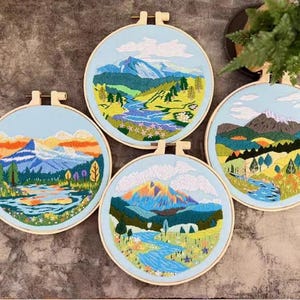 May include: Four round embroidery hoops with landscape scenes. Each hoop features a different mountain range, river, and foliage design. The embroidery uses various shades of blue, green, yellow, and orange thread, creating a colorful and detailed artwork.
