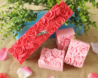 May include: Handmade soaps featuring a rose design. The soaps are in shades of pink and red, with a rectangular bar and three square bars. The soaps are decorated with detailed rose patterns.