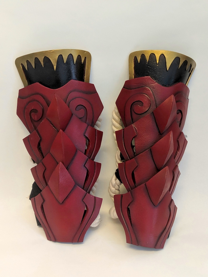 May include: A pair of red leather arm guards with gold and black accents. The guards are decorated with a pattern of scales and swirls.