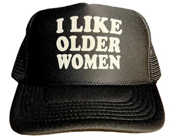 Peut inclure: Un chapeau de camionneur noir avec un filet blanc à l'arrière. Le devant du chapeau porte le texte "I LIKE OLDER WOMEN" en blanc.