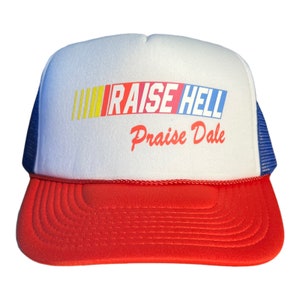 Pode incluir: Um chapéu de caminhoneiro branco e vermelho com uma malha azul nas costas. A frente do chapéu tem um gráfico colorido que diz "RAISE HELL" com as palavras "Praise Dale" abaixo.