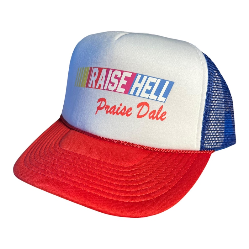 Op de afbeelding: Een witte en rode truckercap met een blauw mesh-achterpaneel. De cap heeft de tekst "RAISE HELL Praise Dale" in kleurrijke letters op de voorkant gedrukt.
