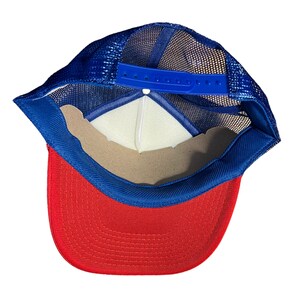 May include: A red and blue mesh trucker hat with a blue adjustable strap. The hat has a red brim and a blue mesh crown.