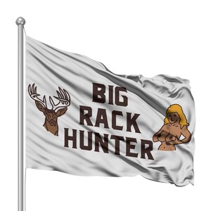 Puede incluir: Una bandera blanca con el texto "BIG RACK HUNTER" en letras marrones. Una cabeza de ciervo marrón con cuernos está en el lado izquierdo de la bandera. Una imagen de dibujos animados de una mujer con cabello rubio está en el lado derecho de la bandera.
