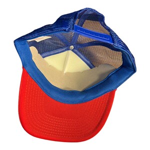 Op de afbeelding: Een rode en blauwe trucker-pet met een mesh-rug. De pet heeft een blauwe verstelbare band en een bruin zweetband.