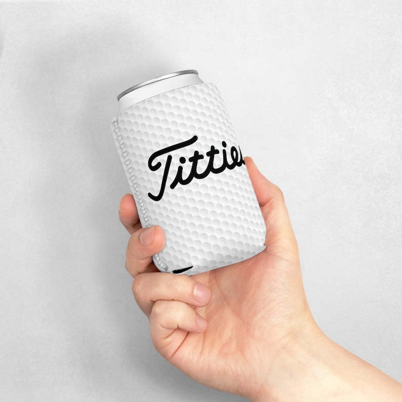 May include: White can cooler with a golf ball pattern and the word "Titties" in black cursive lettering.