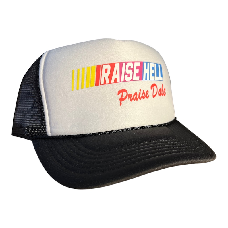 Op de afbeelding: Een witte truckercap met een zwart mesh achterpaneel. De voorkant van de cap heeft een kleurrijk race-streepontwerp met de tekst "RAISE HELL Praise Dale" in rood en wit.