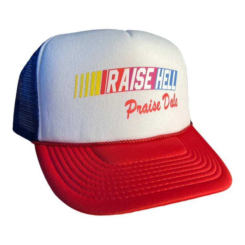 Op de afbeelding: Een witte en rode trucker-pet met een blauw mesh-achterpaneel. De voorkant van de pet heeft een kleurrijk race-streepontwerp en de tekst "RAISE HELL Praise Dale".