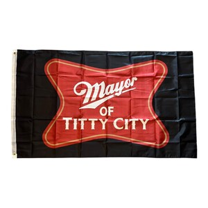 Może przedstawiać: Czarna flaga z czerwonym i złotym emblematem z napisem "Mayor of Titty City" w białym skrypcie. Flaga ma oczka do zawieszania.