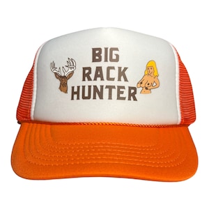 May include: A white and orange trucker hat with the text "BIG RACK HUNTER" in brown letters. A cartoon deer and a woman are on the hat.