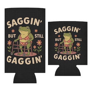 May include: Black can cooler with a humorous graphic of a frog in a polka dot bikini using a walker. The text reads "Saggin' But Still Gaggin'" with flower accents. A smaller version is also shown.