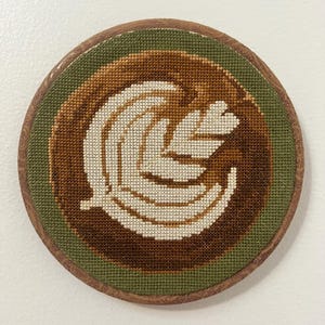 May include: A circular cross-stitch artwork showing a coffee latte art design. The design features a leaf pattern in cream and brown shades, set against a brown and green background, framed by a wooden border.