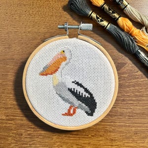 May include: A cross-stitch embroidery hoop featuring a pelican design. The pelican is depicted with a white body, black wing details, and an orange beak and feet. The embroidery is set in a wooden hoop, with colorful embroidery floss in the background.