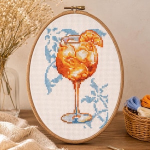 Op de afbeelding: Een ovale houten borduurring met een kruissteekpatroon van een oranje cocktail in een glas op voet. Het ontwerp toont een feloranje drankje met ijsblokjes, een schijfje citrusvrucht als garnering en lichtblauwe decoratieve bloemaccenten.