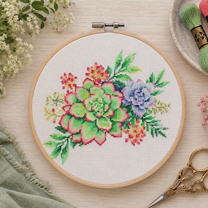 May include: A circular wooden embroidery hoop containing white Aida fabric with a cross-stitch design of a succulent arrangement. The pattern features green, pink, and purple succulents with small yellow accents and green leaves on a white grid.