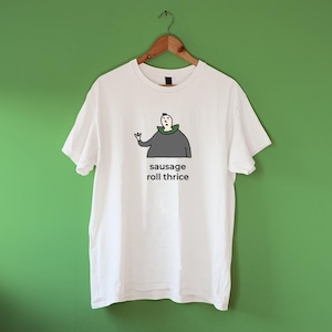 Op de afbeelding: Wit T-shirt met een cartoonillustratie van een persoon die een groene capuchon draagt en de tekst "sausage roll thrice".