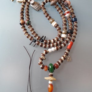 May include: A brown and white beaded necklace with various charms, including a green stone, a red coral bead, and a silver charm. The necklace is strung on a brown cord.