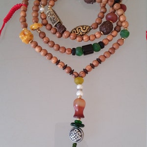 May include: A brown wooden mala bead necklace with various charms, including a green jade bead, a gold-colored dragon charm, and a silver knot charm. The necklace has a green tassel.